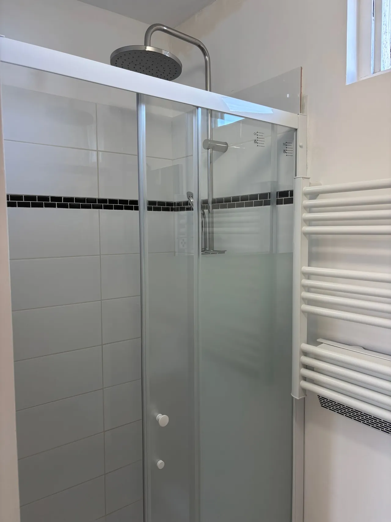 Douche terminée : porte coulissante vitrée, colonne de douche à pommeau, faïence blanche à frise noire, sèche-serviettes