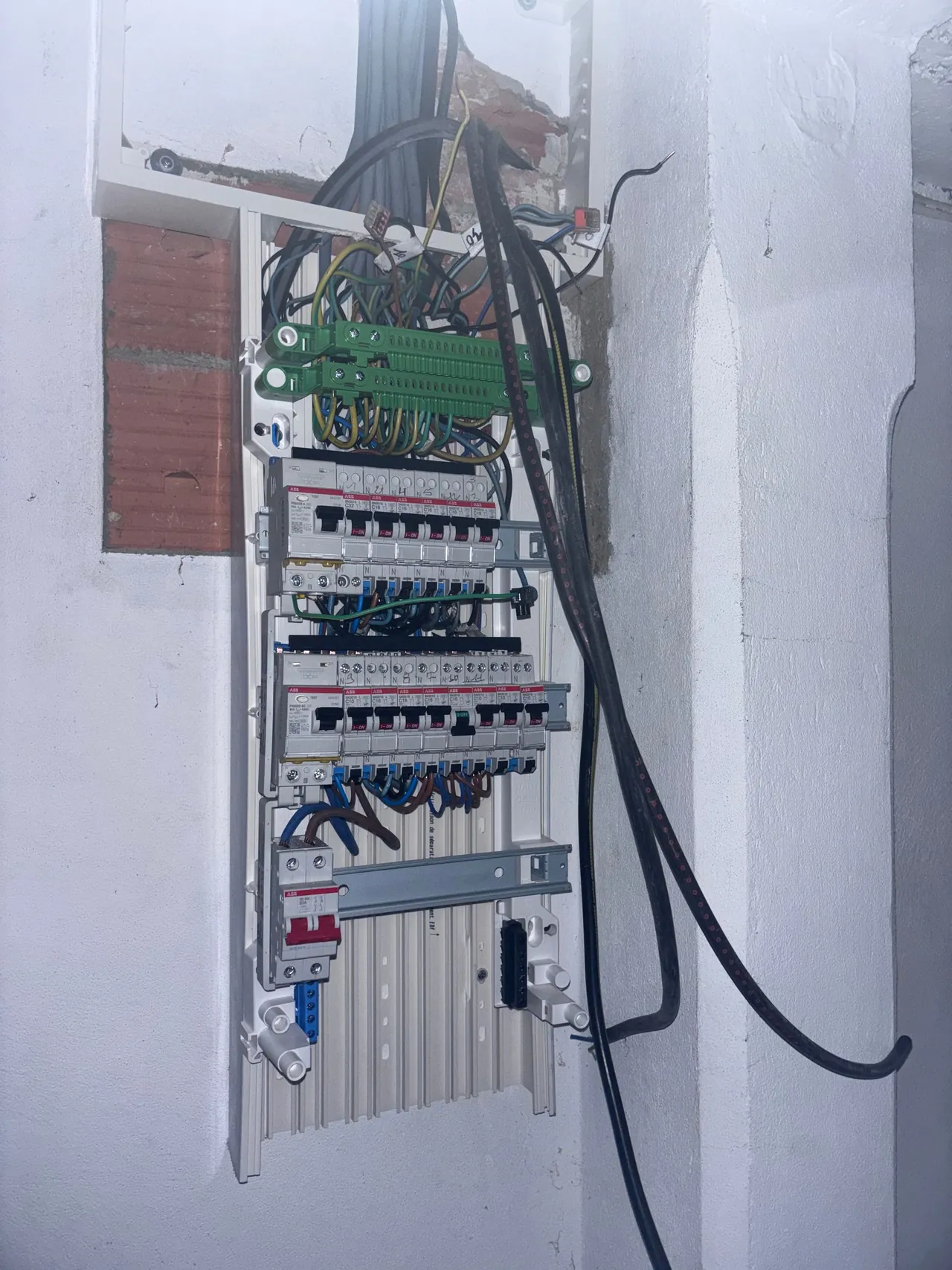 Tableau électrique en cours de câblage dans une niche en briques : rangées de disjoncteurs, peignes, borniers verts et câbles d’arrivée